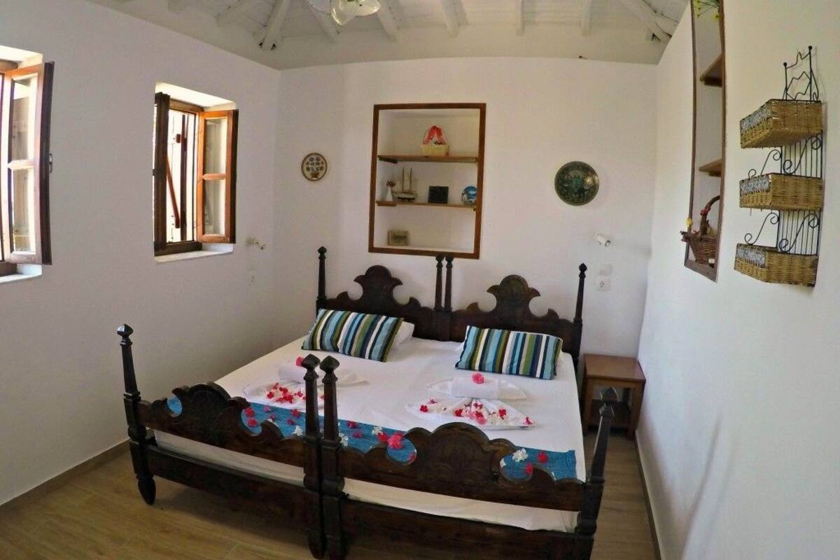 A bedroom with a wooden four-poster bed, a small bedside table, a wall-mounted shelf, and a window with curtains.