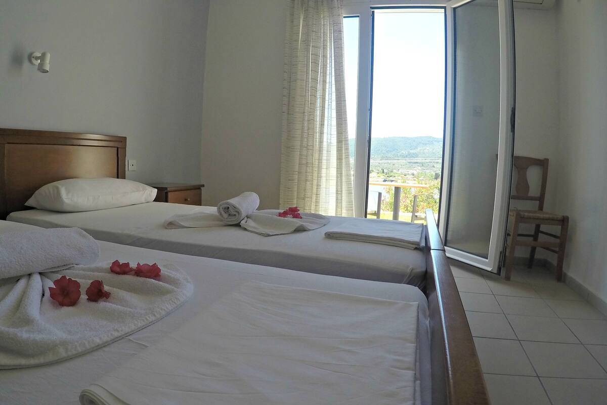 A hotel room with two beds, each with neatly folded towels and a flower. There is a wooden chair and a small table by the window.