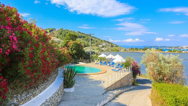 A pool area with lounge chairs and umbrellas, surrounded by greenery and a view of the water. - Villa Ariadni (Skiathos)