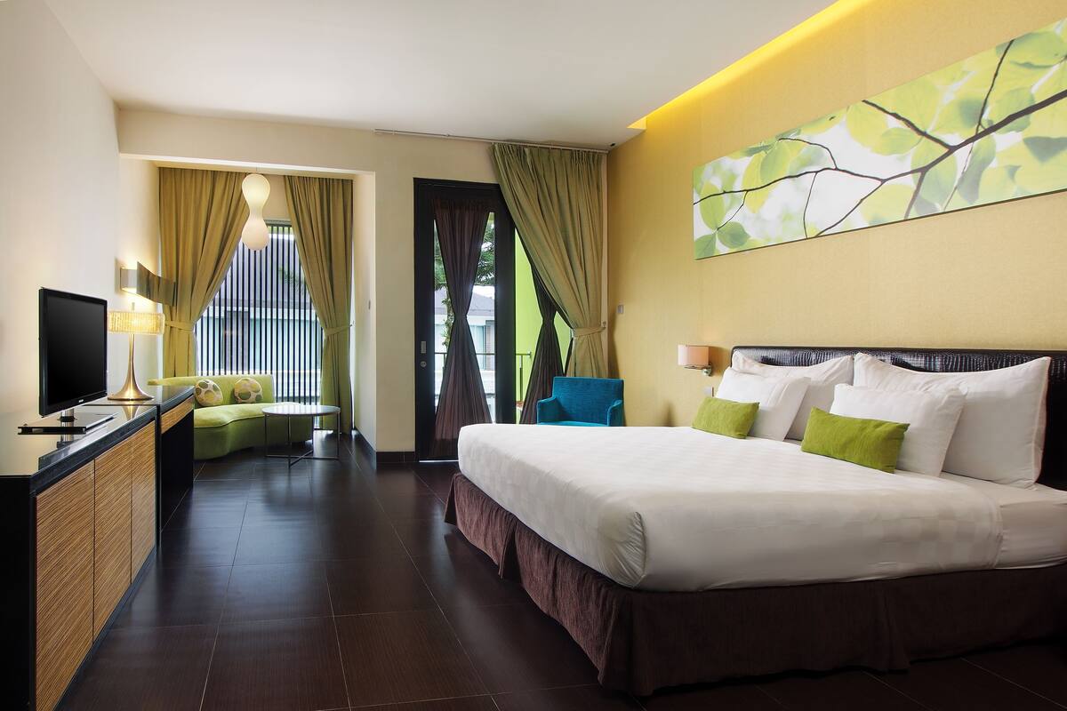 Een hotelkamer met een groot bed, een bureau met een flatscreen-televisie, een bank en een stoel. Er is een raam met gordijnen en een uitzicht op groen.