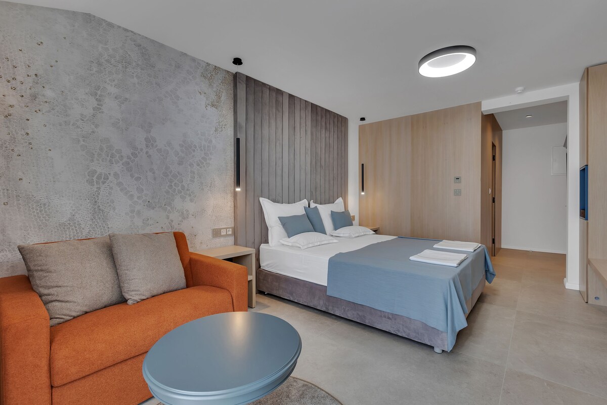 A modern hotel room with a bed, a sofa, a round table, and a large window.