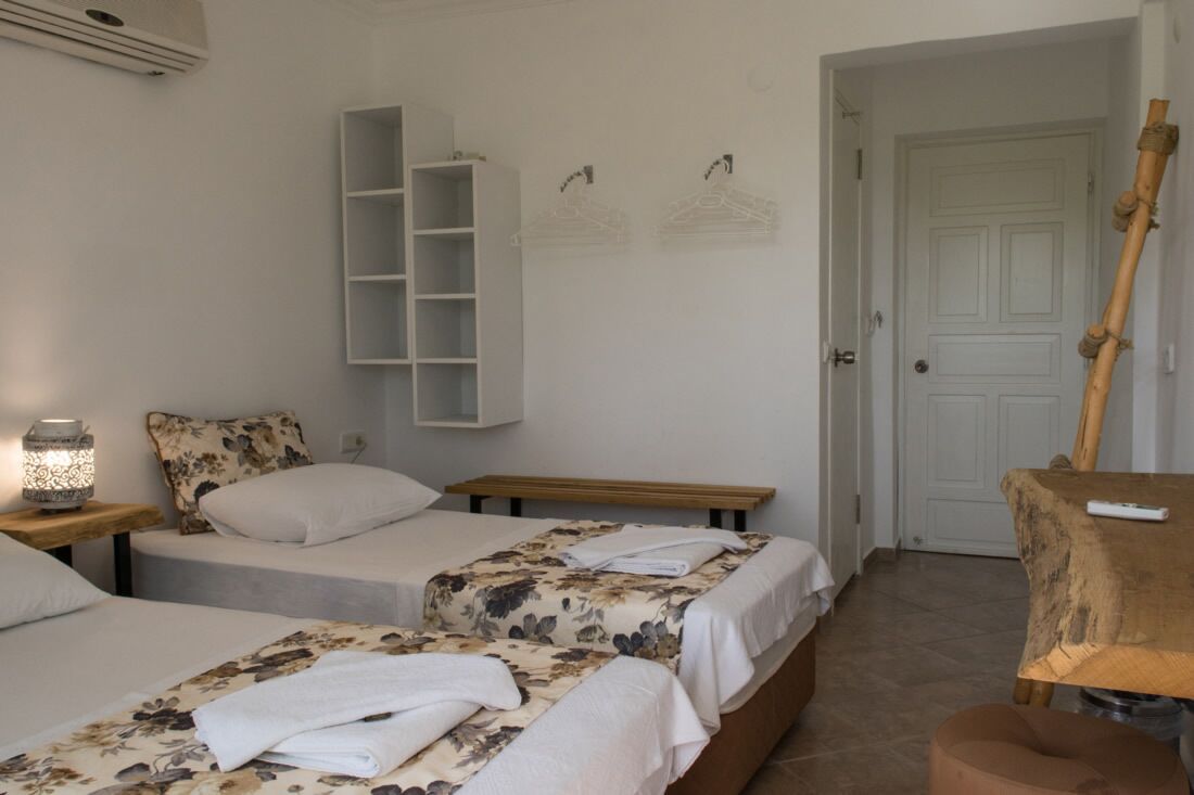 A hotel room with two beds, a bedside table with a lamp, a wardrobe, and a door leading to another room.