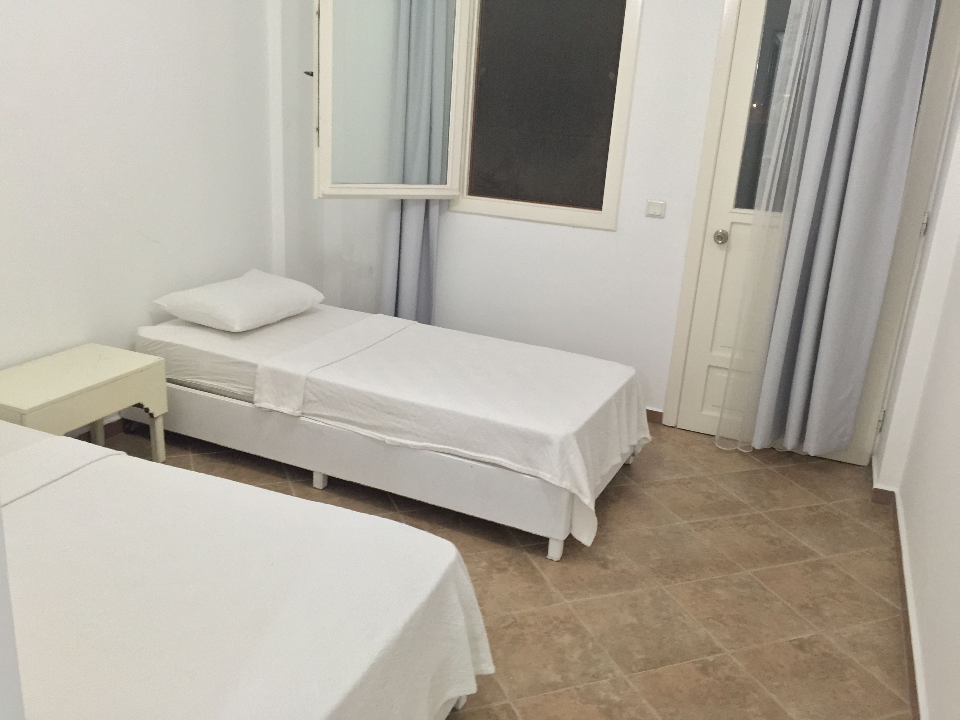Quarto pequeno e limpo com duas camas de solteiro, uma mesa de cabeceira, janela com cortinas e porta.