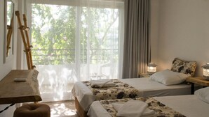 A hotel room with two beds, a wooden table, a chair, and a balcony with a view of trees. - Hotel Pataros (Kas)