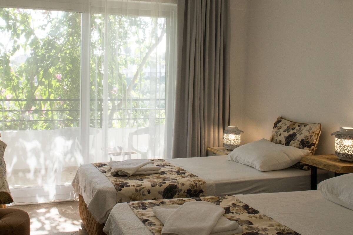A room with two beds, a wooden table, a chair, a lamp, and a window with a view of greenery.