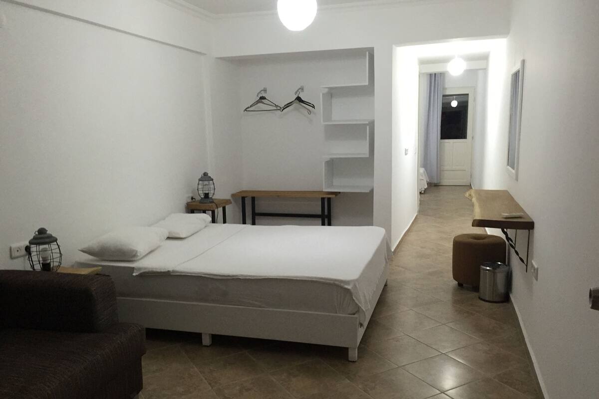 A bedroom with a bed, a bedside table, a lamp, a hanging light fixture, a bench, a trash can, and a small stool.