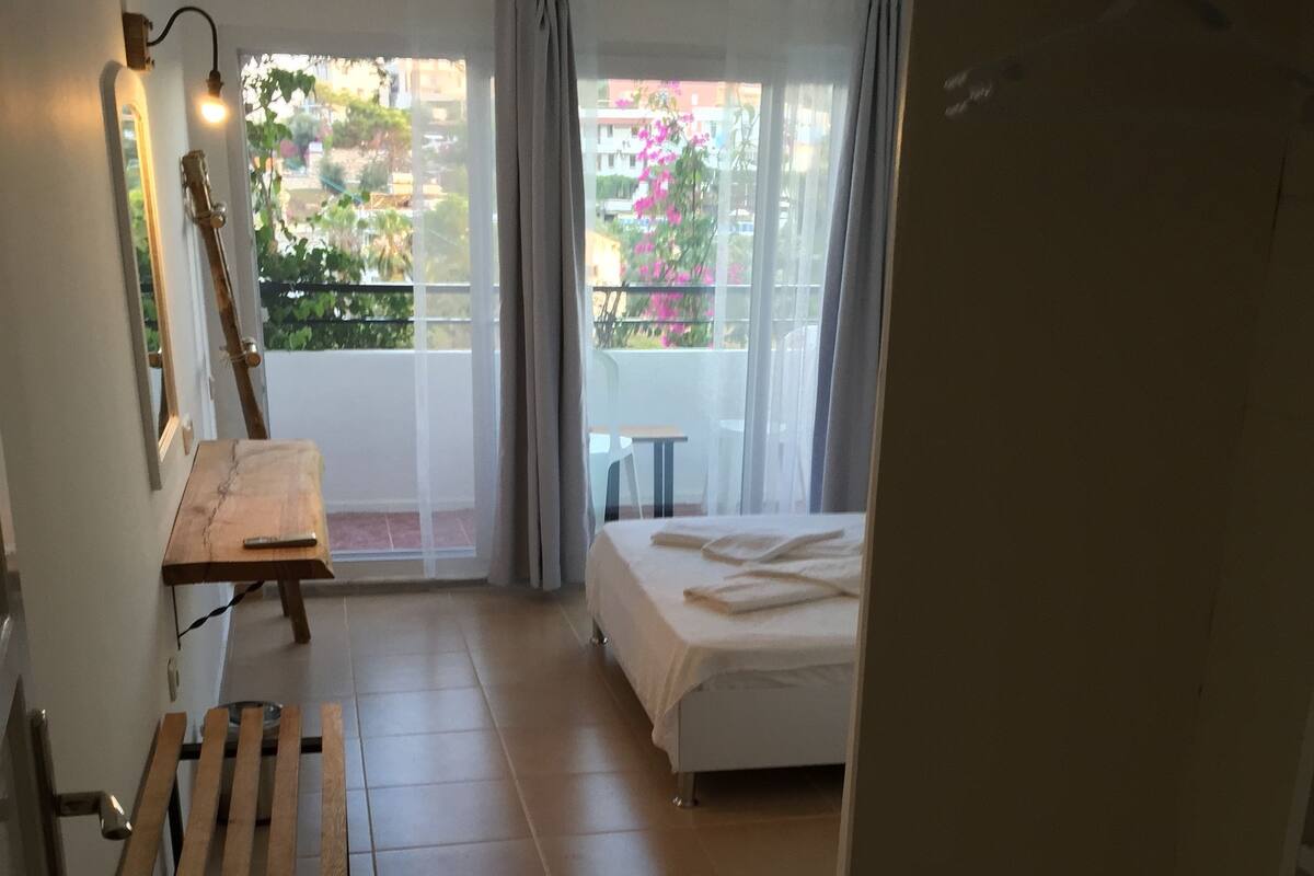 A room with a bed, a small table, a bench, and a view of a balcony with plants and a building.