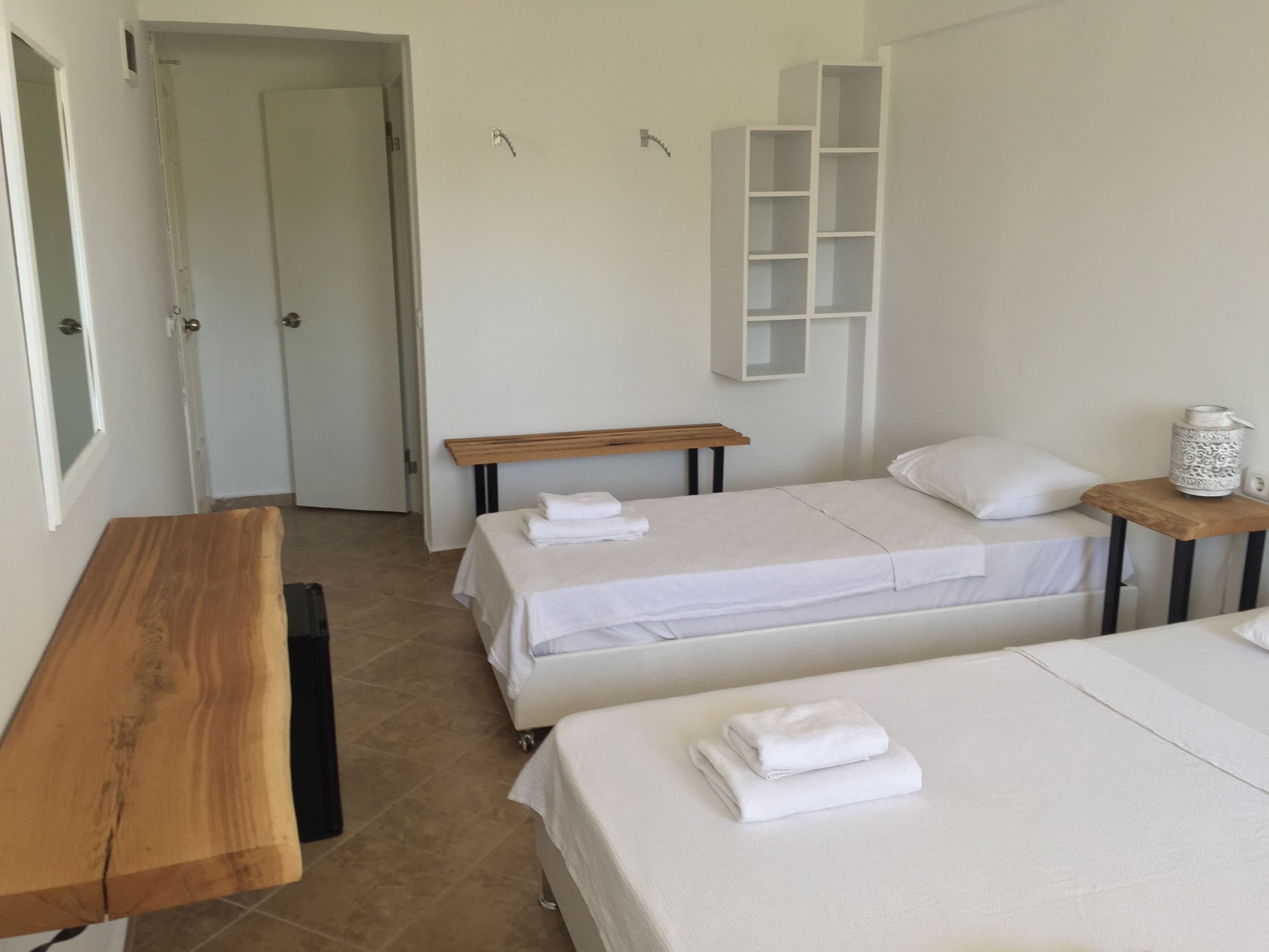 Une chambre d’hôtel avec deux lits, un banc en bois, une petite table et une étagère blanche.