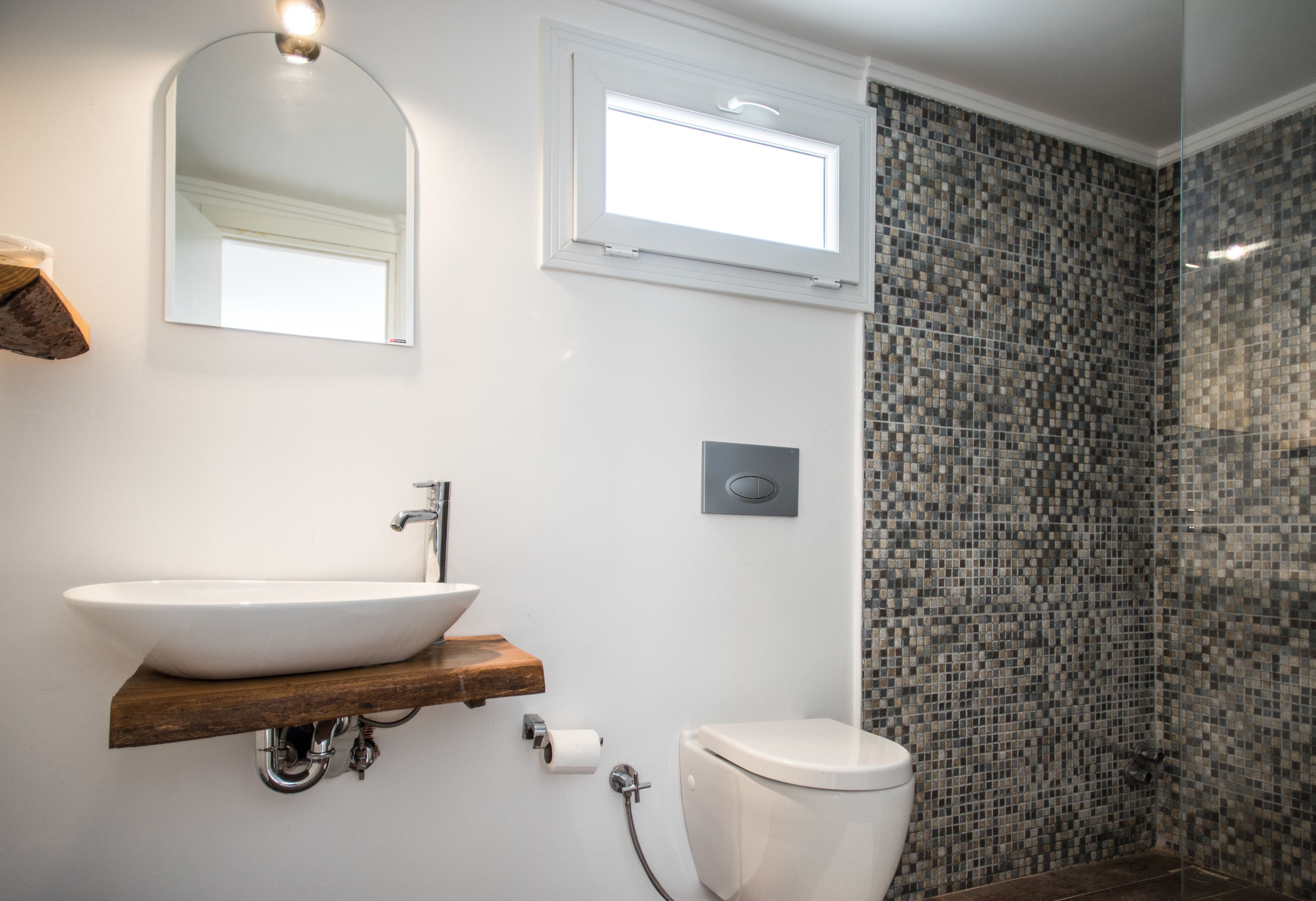 Ein Badezimmer mit Waschbecken, Toilette, Dusche mit Mosaikfliesen, Holzregal und Spiegel.