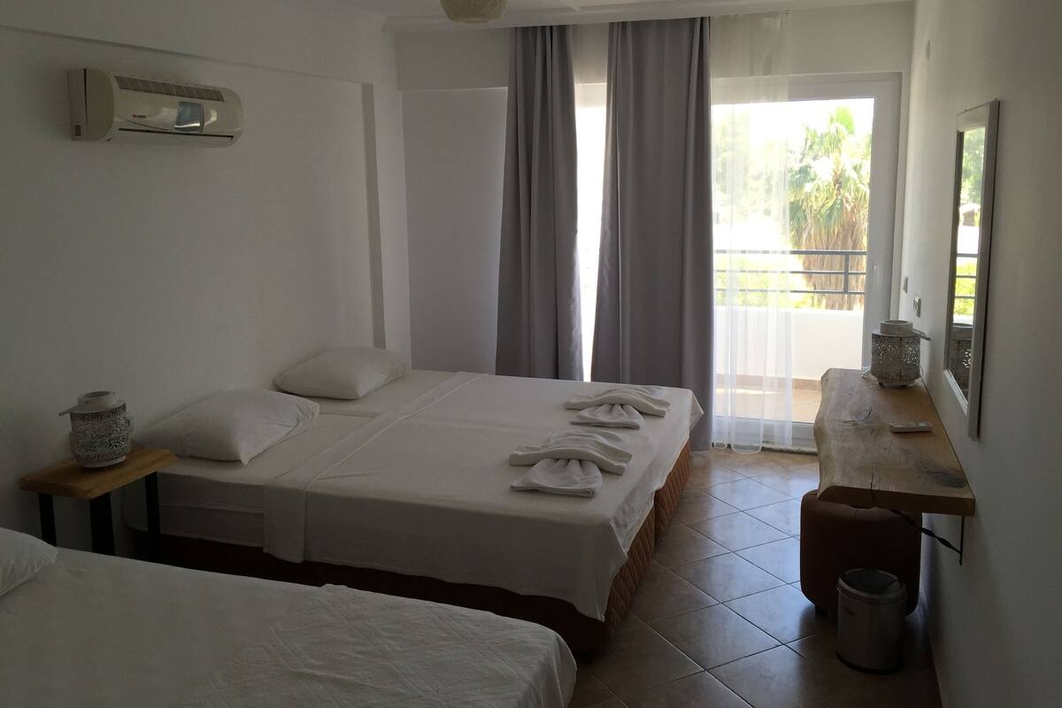 A hotel room with two beds, a small table, a trash bin, and a balcony with curtains.
