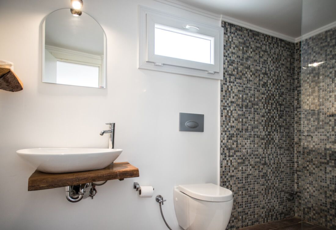 A bathroom with a white sink, a toilet, a shower with mosaic tiles, a wooden shelf, and a mirror.
