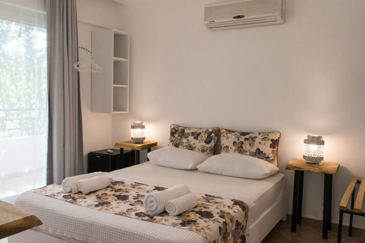 A neatly made bed with floral bedding, a bedside table with a lamp, a wall-mounted air conditioner, and a window with curtains.
