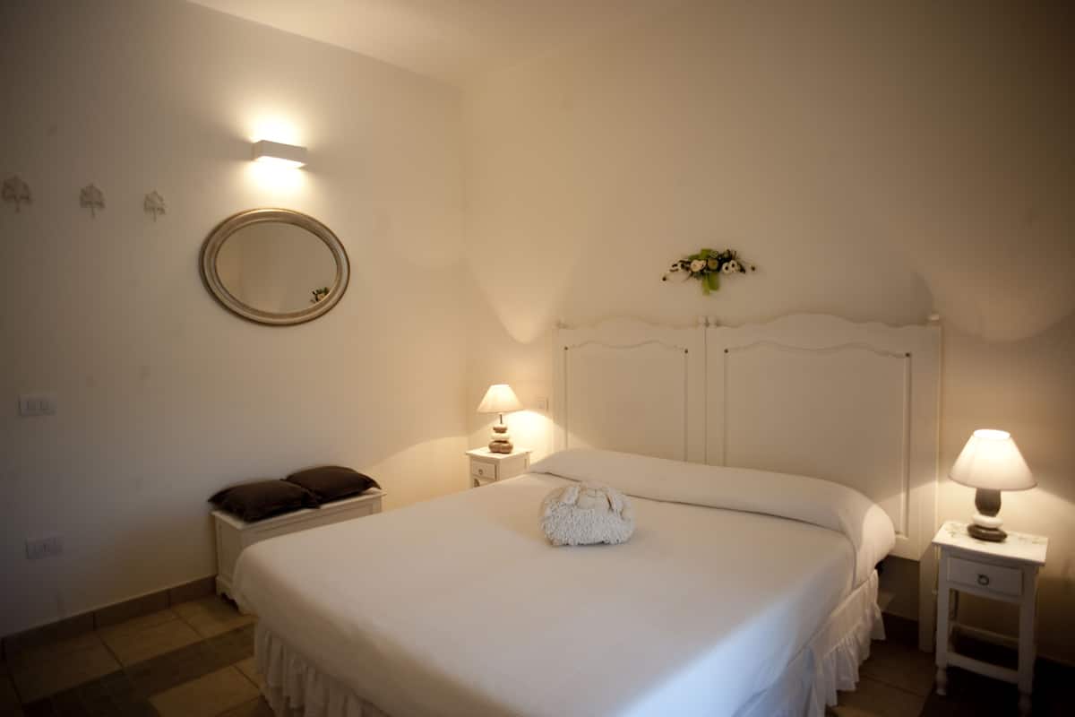 A neatly made bed with a white comforter and a fluffy white towel in the center. There is a round mirror on the wall above the bed and a small bedside table with a lamp.