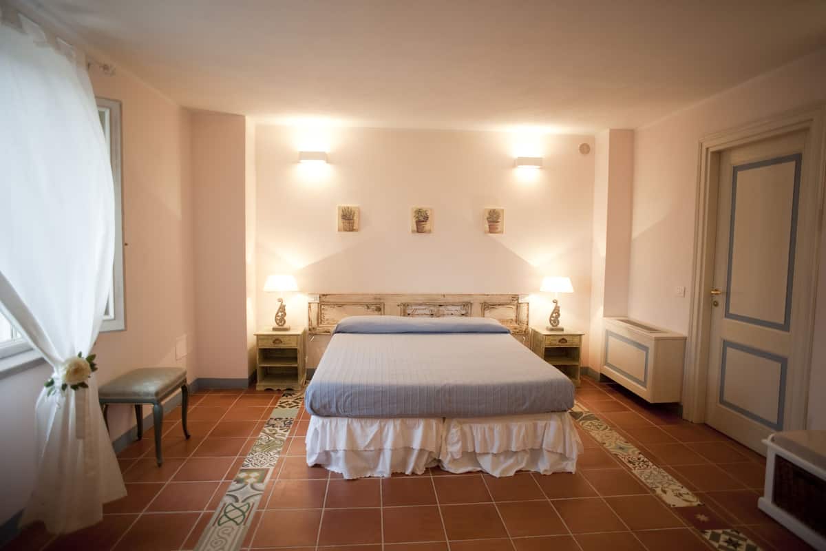 A bedroom with a bed, bedside lamps, a bench, a door, and a wall with three potted plants.