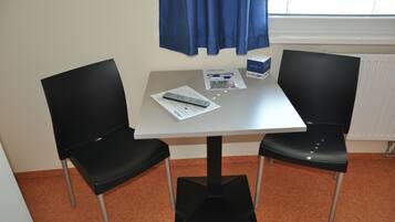 Una habitación pequeña con una mesa blanca, dos sillas negras, un ventanal con cortinas azules y un radiador.