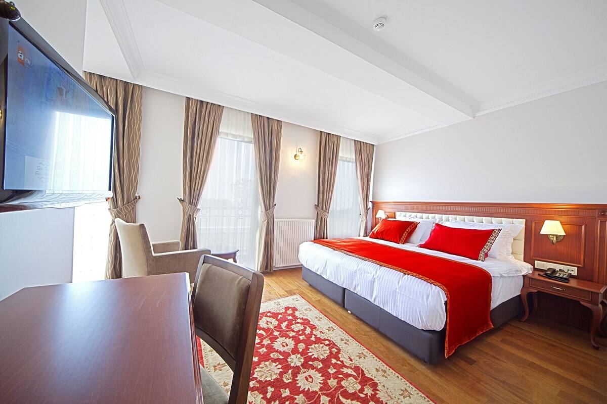 A hotel room with a large bed, a desk, a chair, a TV, and a window with curtains.