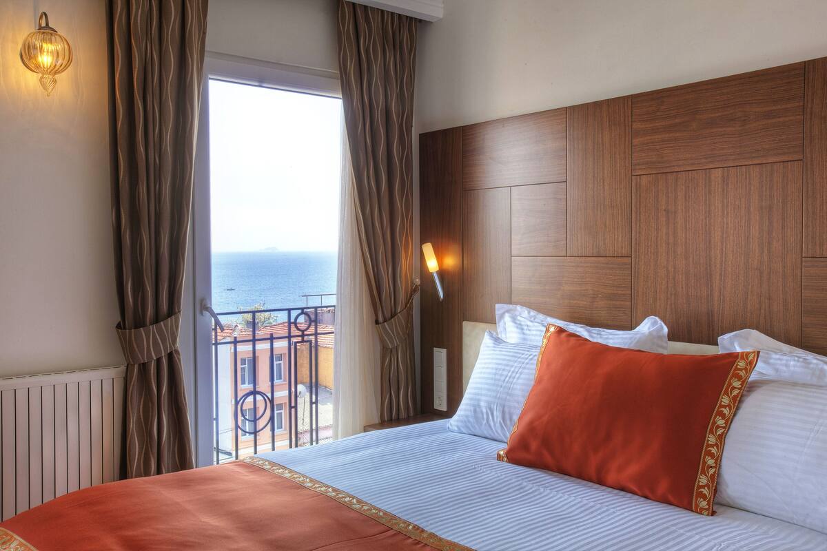 A hotel room with a large bed, a wooden headboard, a view of the sea, and a balcony with a railing.