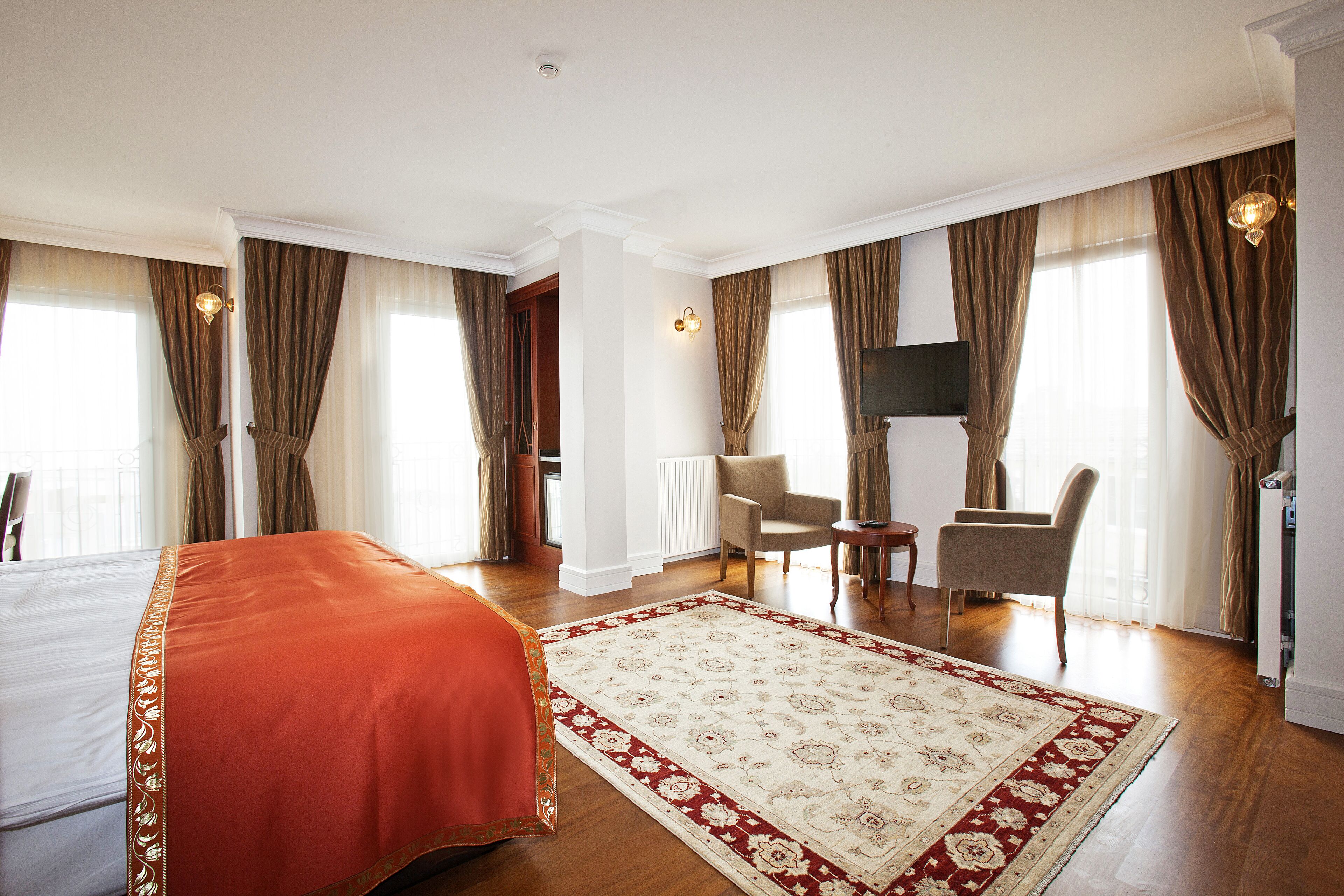 Habitación de hotel con una cama grande, zona de estar con dos sillones, televisión y un ventanal con cortinas.