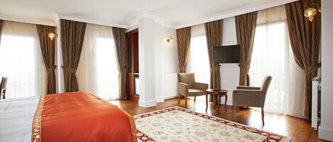 Habitación de hotel con una cama grande, zona de estar con dos sillones, televisión y un ventanal con cortinas.
