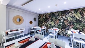 A modern dining area with white tables and chairs, a tropical leaf-patterned wall, and decorative wall art.