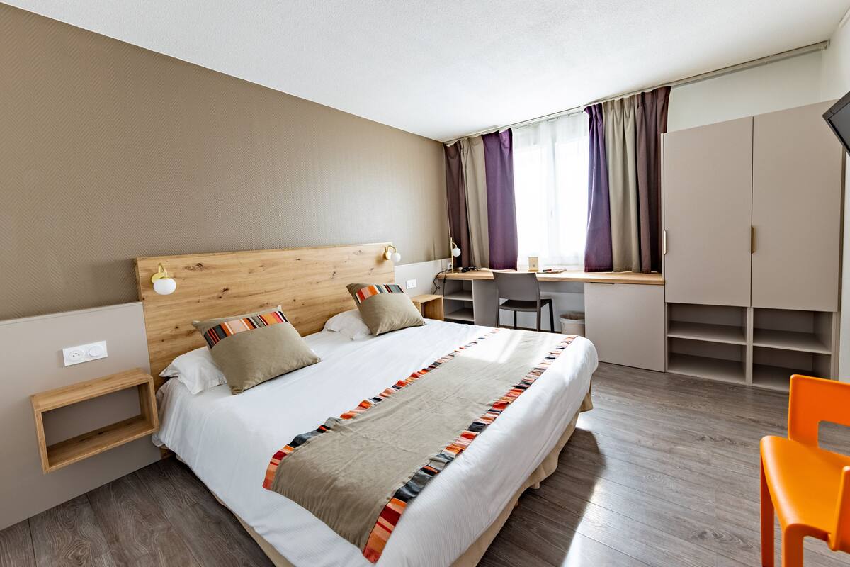 Una habitación de hotel moderna con cabecera de madera, una cama grande con manta colorida, un escritorio con silla y un armario.