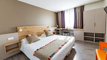 A modern hotel room with a wooden headboard, a large bed with a colorful blanket, a desk with a chair, and a wardrobe.