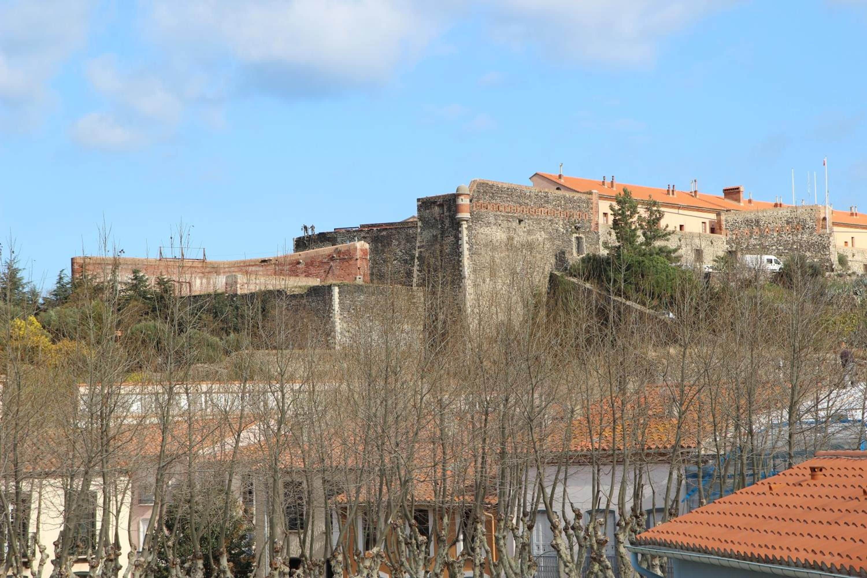 A historic stone building with a red-tiled roof, surrounded by leafless trees and a clear sky.