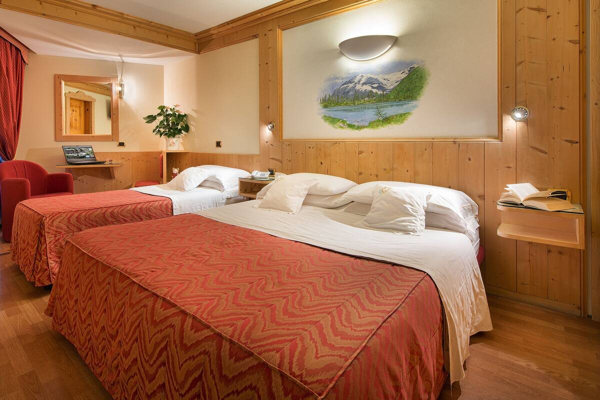 Habitación de hotel con dos camas, paredes de madera, un cuadro de un paisaje montañoso y un escritorio con una computadora.