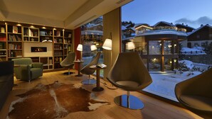 A modern living room with a large bookshelf, comfortable seating, and a view of a snowy landscape through large windows. - Hotel Baita Montana (Livigno)