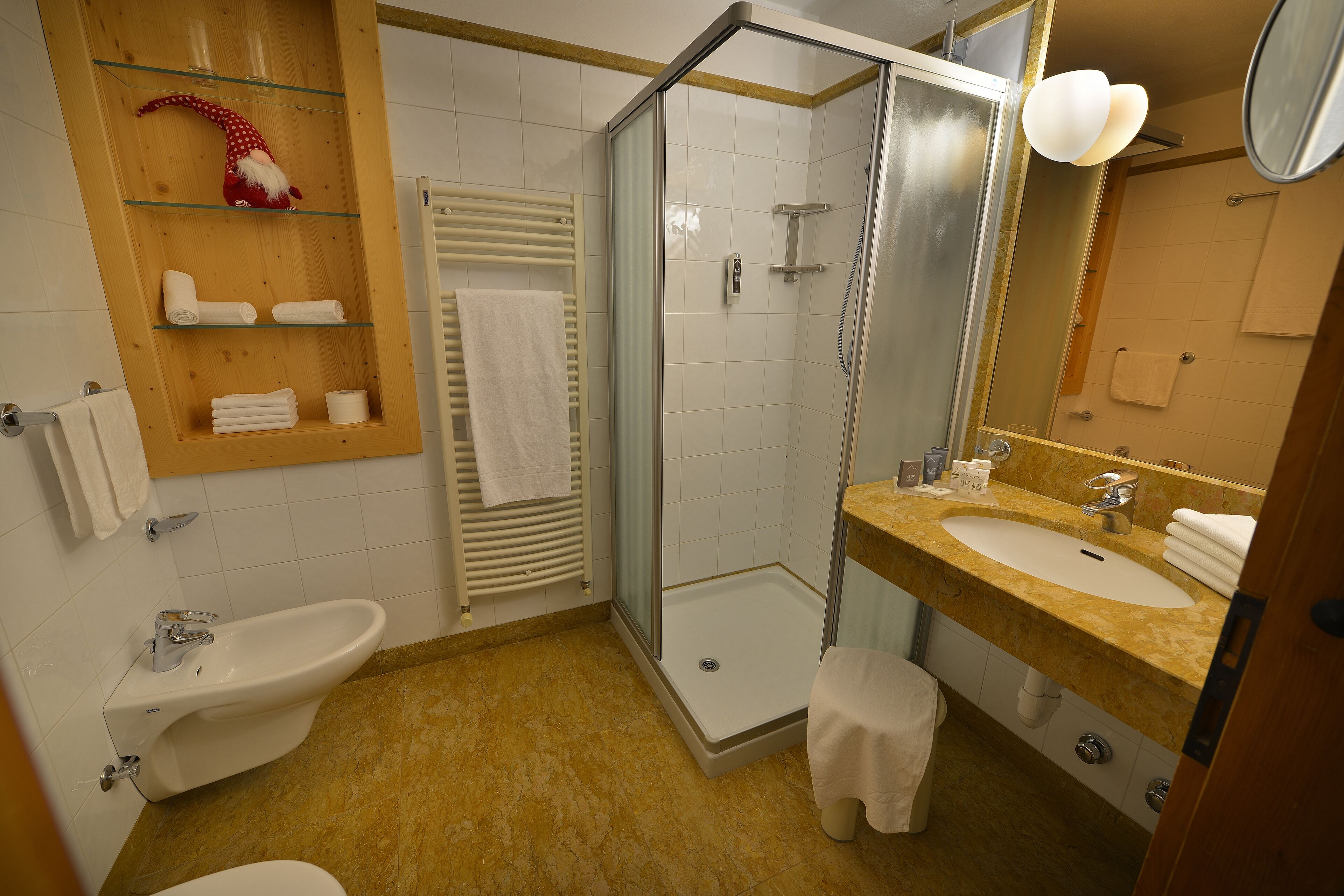 A bathroom with a glass-enclosed shower, a sink with a marble countertop, a bidet, and a wooden shelf with towels.