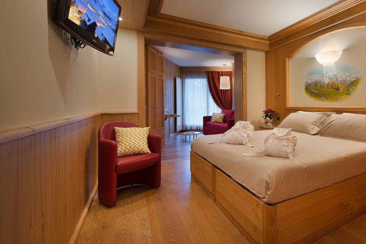 Habitación de hotel con una cama de madera, un sillón rojo, una mesita con una lámpara y un cuadro en la pared.