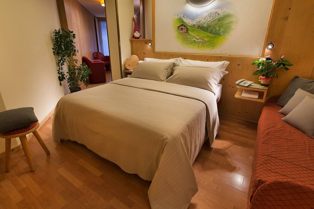 Habitación de hotel con una cama, un sofá rojo, suelo de madera, una mesita con una lámpara y un cuadro en la pared.