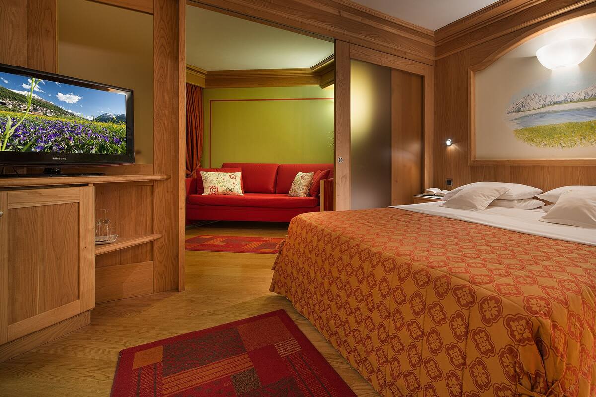 Habitación de hotel con una cama grande, un televisor, un cuadro y un sofá rojo en la habitación contigua.