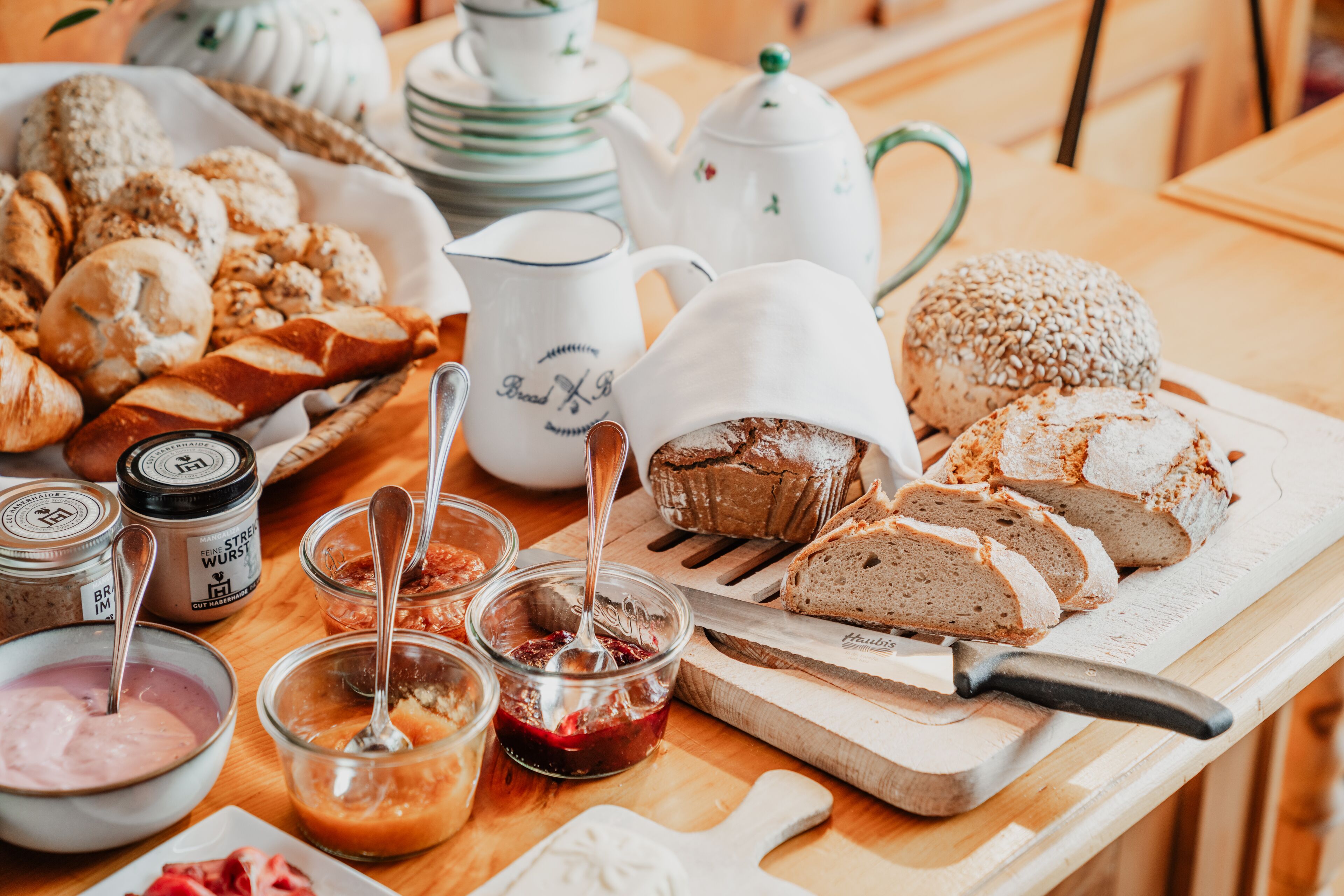 Verschiedene Brotsorten und Gebäck auf einem Holztisch, zusammen mit Gläsern mit Brotaufstrichen und einem Schneidebrett mit Messer.