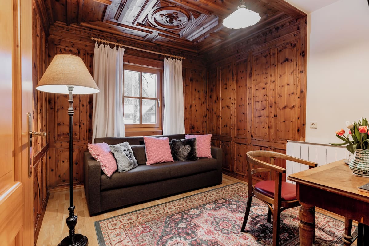 A cozy room with wooden walls, a brown sofa with pink and gray cushions, a wooden dining table with a vase of flowers, and a patterned rug.