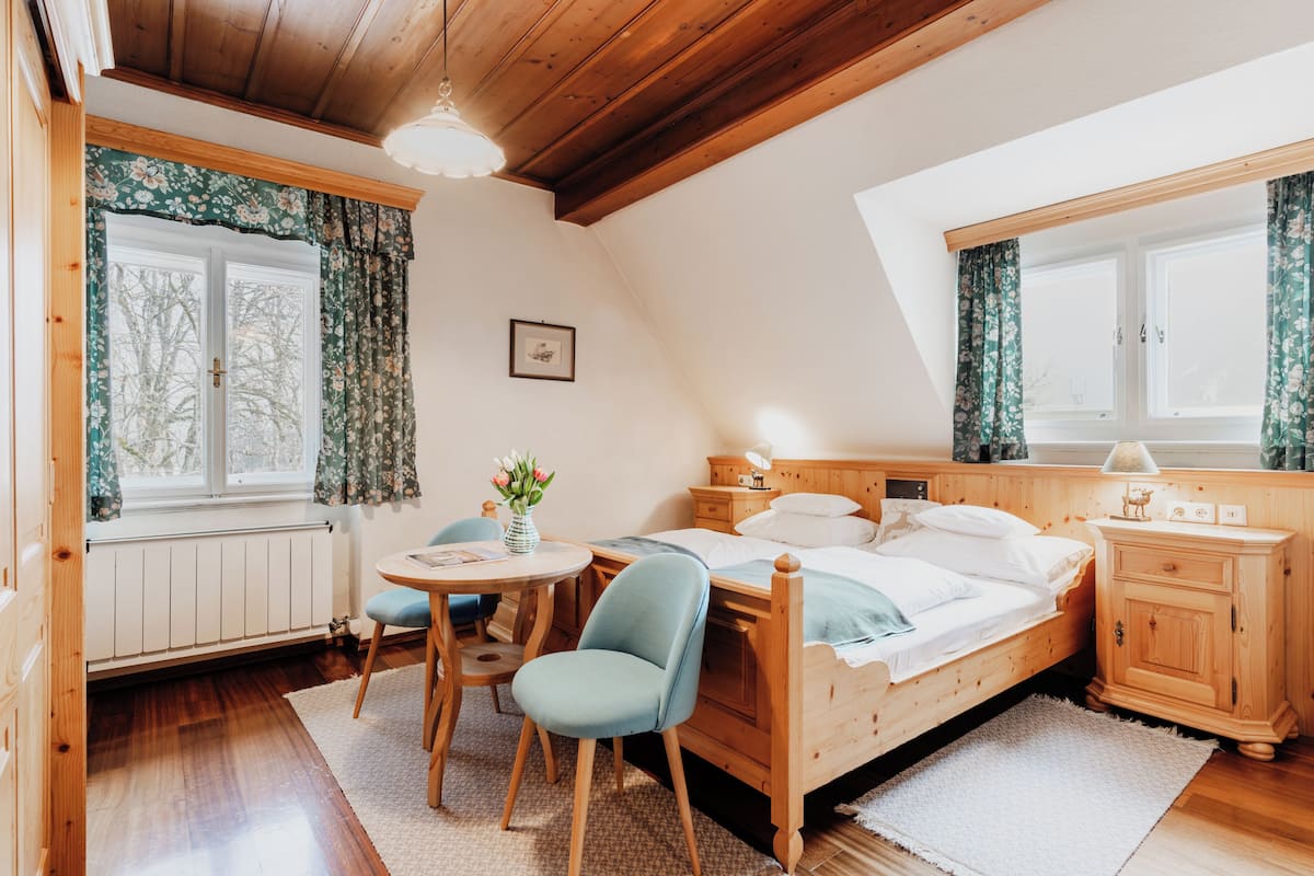 A cozy bedroom with a wooden bed, a bedside table, a chair, a round table, and a window with floral curtains.