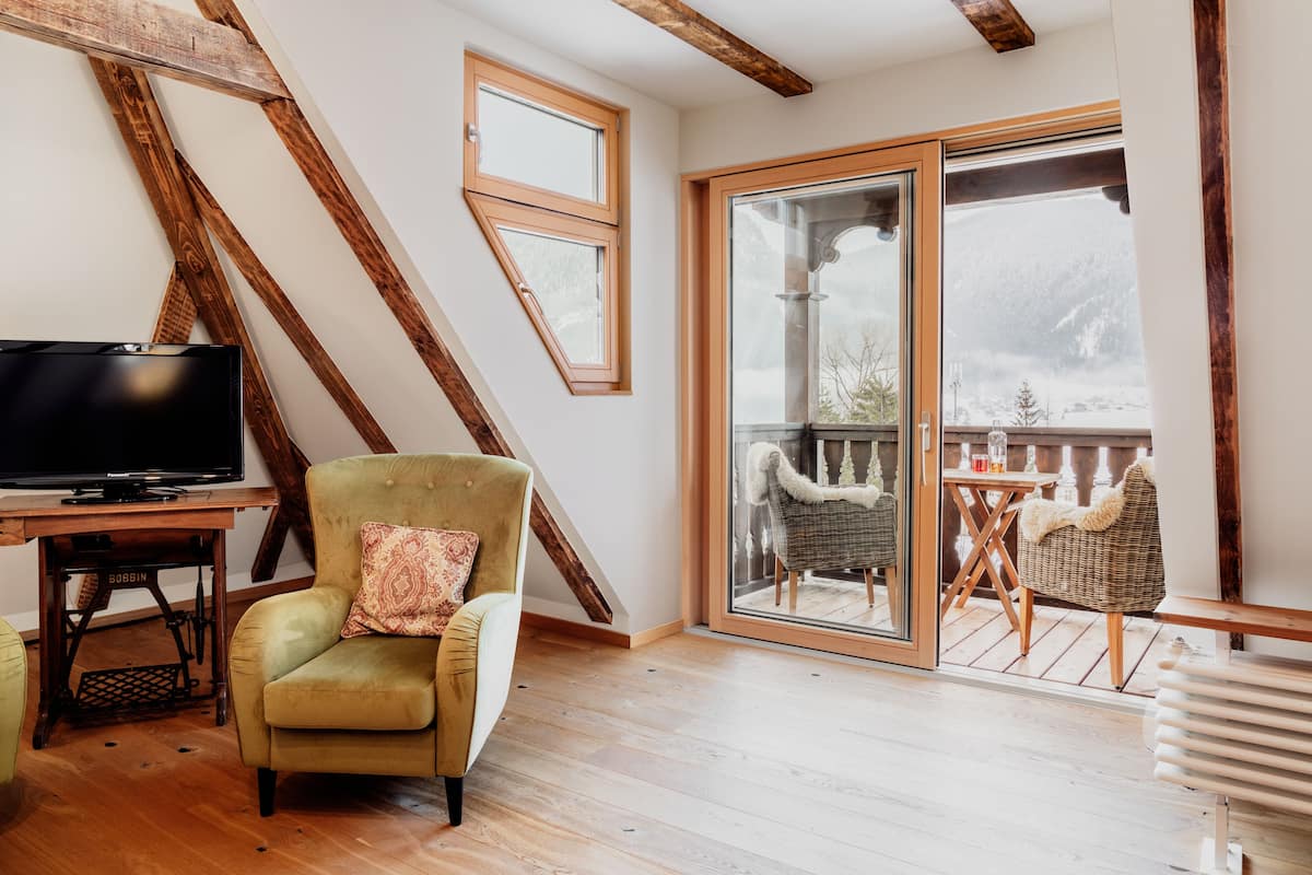 A room with a television, a green armchair, a wooden table, and a balcony with outdoor seating.
