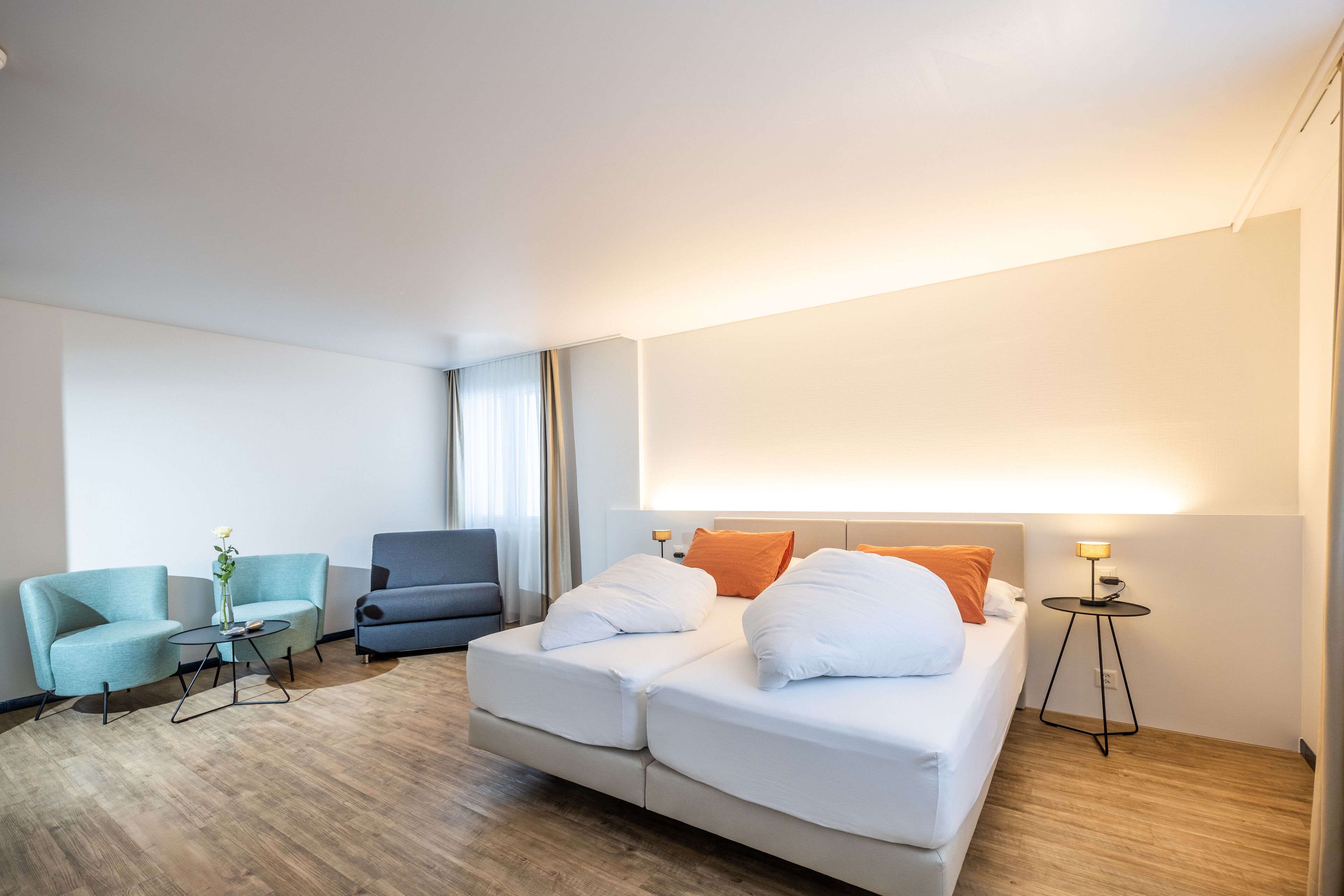 A modern hotel room with a large bed, two armchairs, a small table, and a bedside lamp.