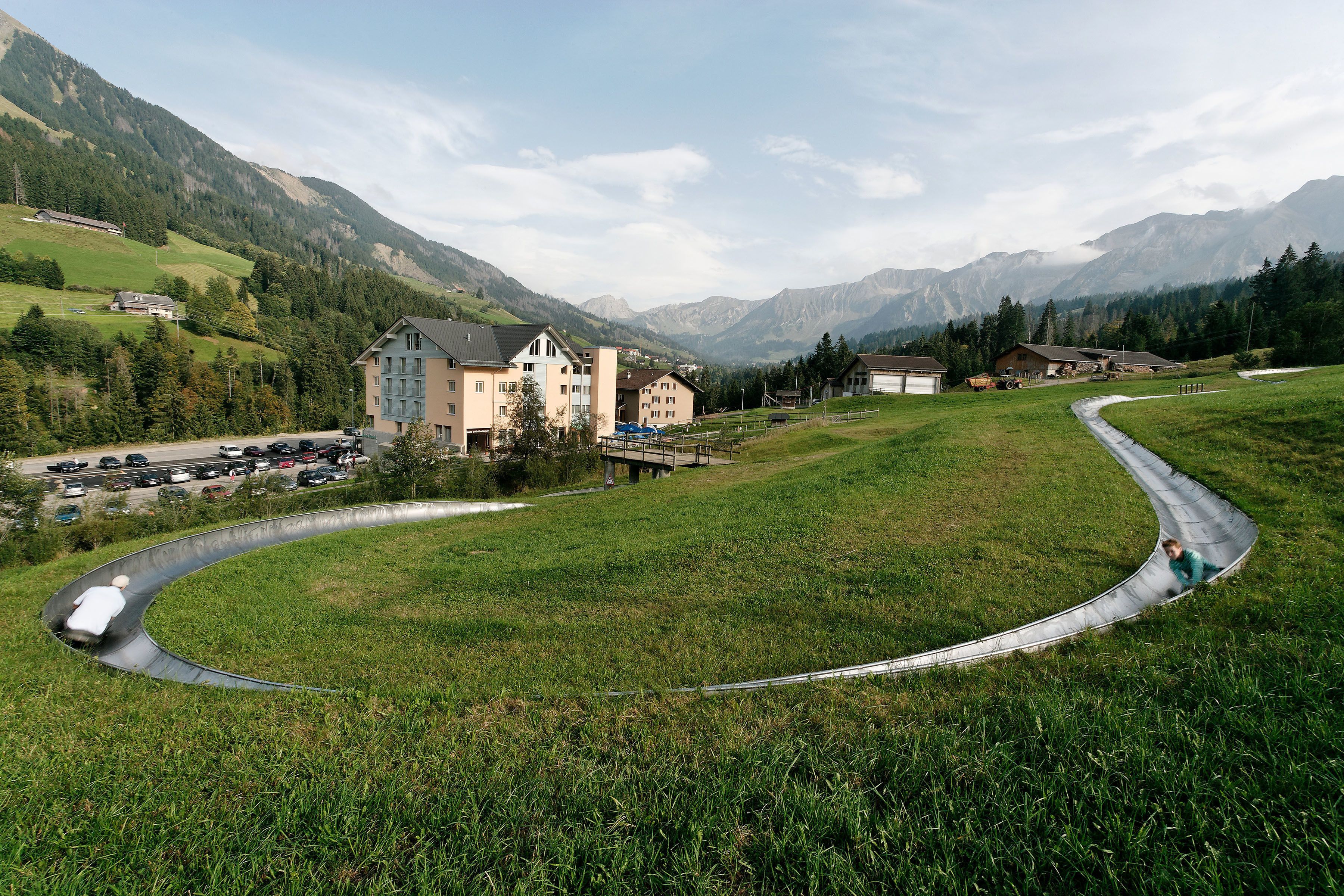 Paesaggio rurale con una strada tortuosa, un edificio ampio con molte finestre e montagne sullo sfondo.
