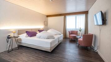 Ein modernes Hotelzimmer mit Bett, Nachttisch, Lampe, Fernseher und einer Sitzecke mit Sesseln.