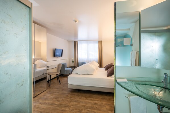A hotel room with a bed, a desk with a chair, a television, a bathroom with a glass sink, and a large mirror.