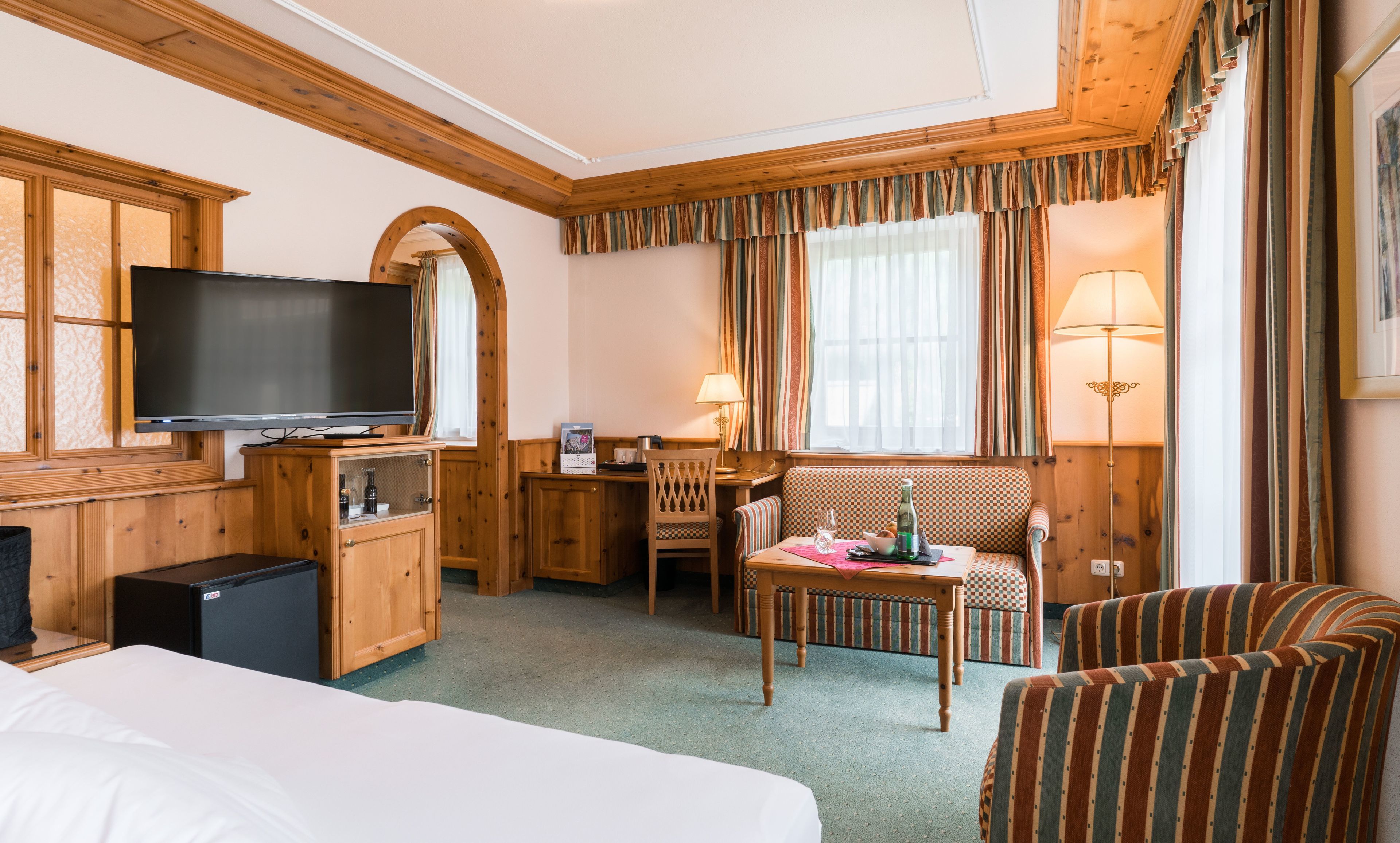 A hotel room with a bed, a TV, a desk with a chair, a small table with a bottle and glasses, and a window with curtains.