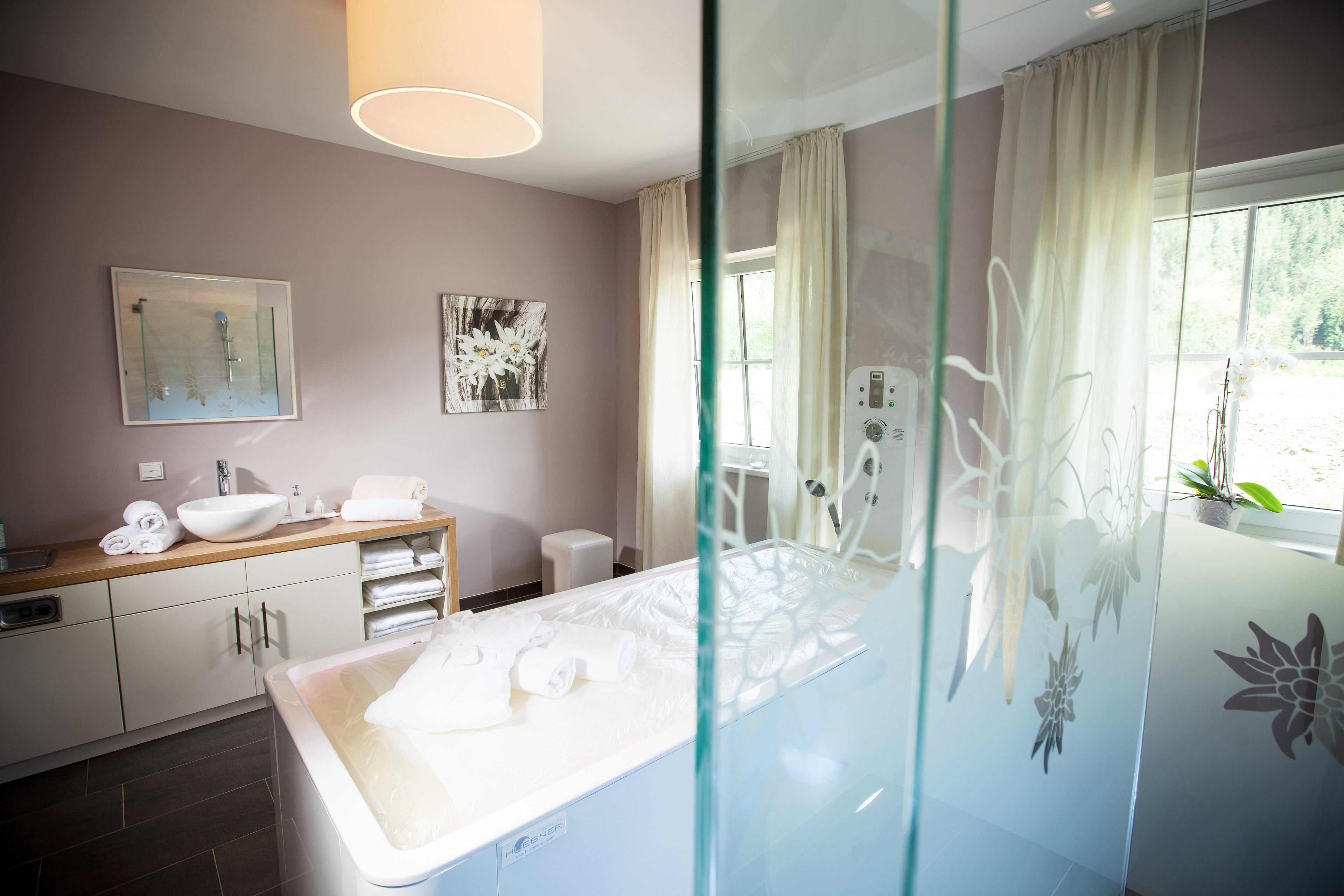 Un bany modern amb plat de dutxa de vidre, un lavabo blanc i una finestra gran amb cortines.