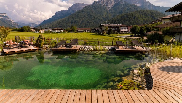 Um lago de montanha límpido com decks de madeira e espreguiçadeiras, emoldurado por uma paisagem montanhosa e edifícios.