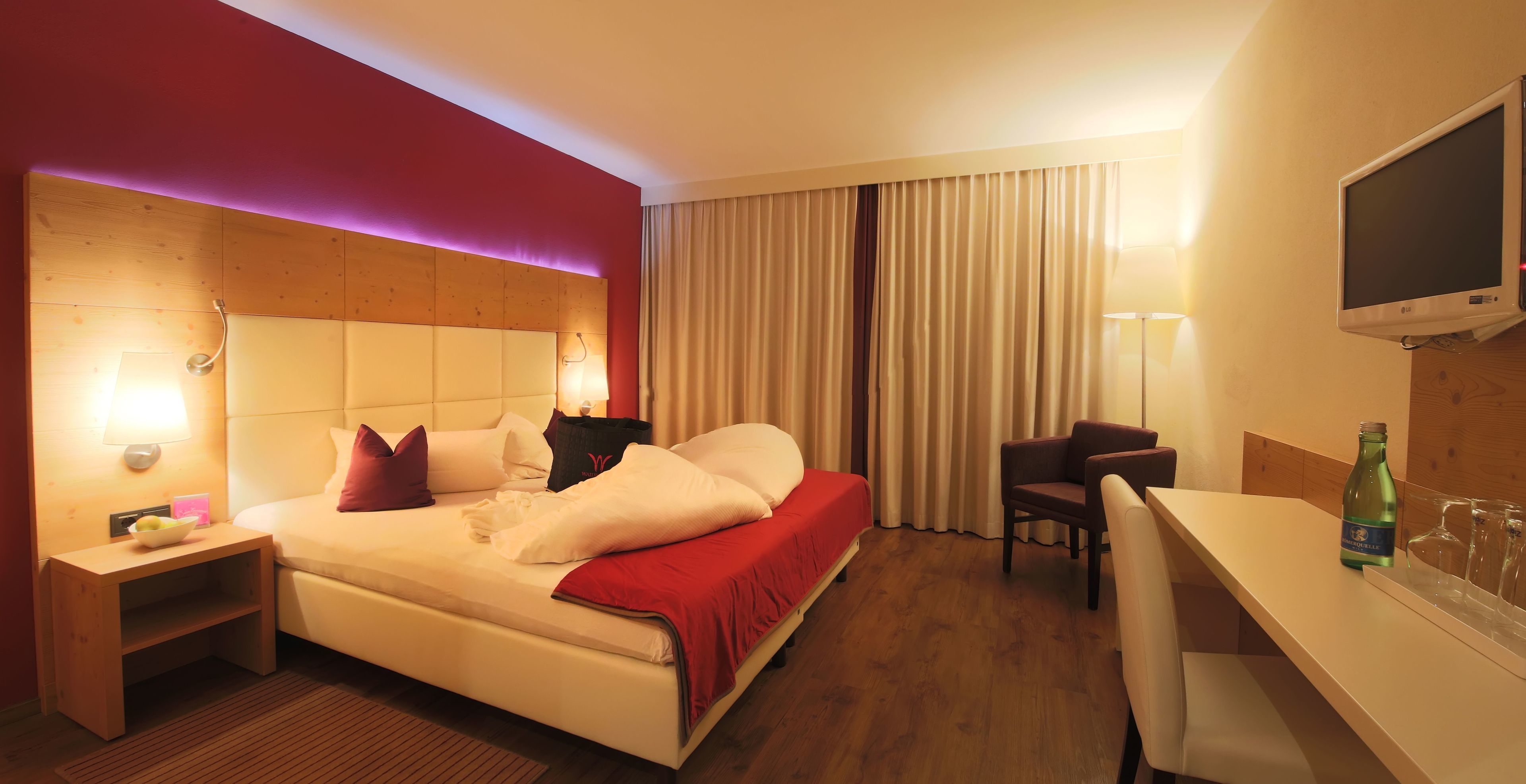 Una habitación de hotel con cama, mesita de noche, silla, televisor y una botella de agua sobre el escritorio.