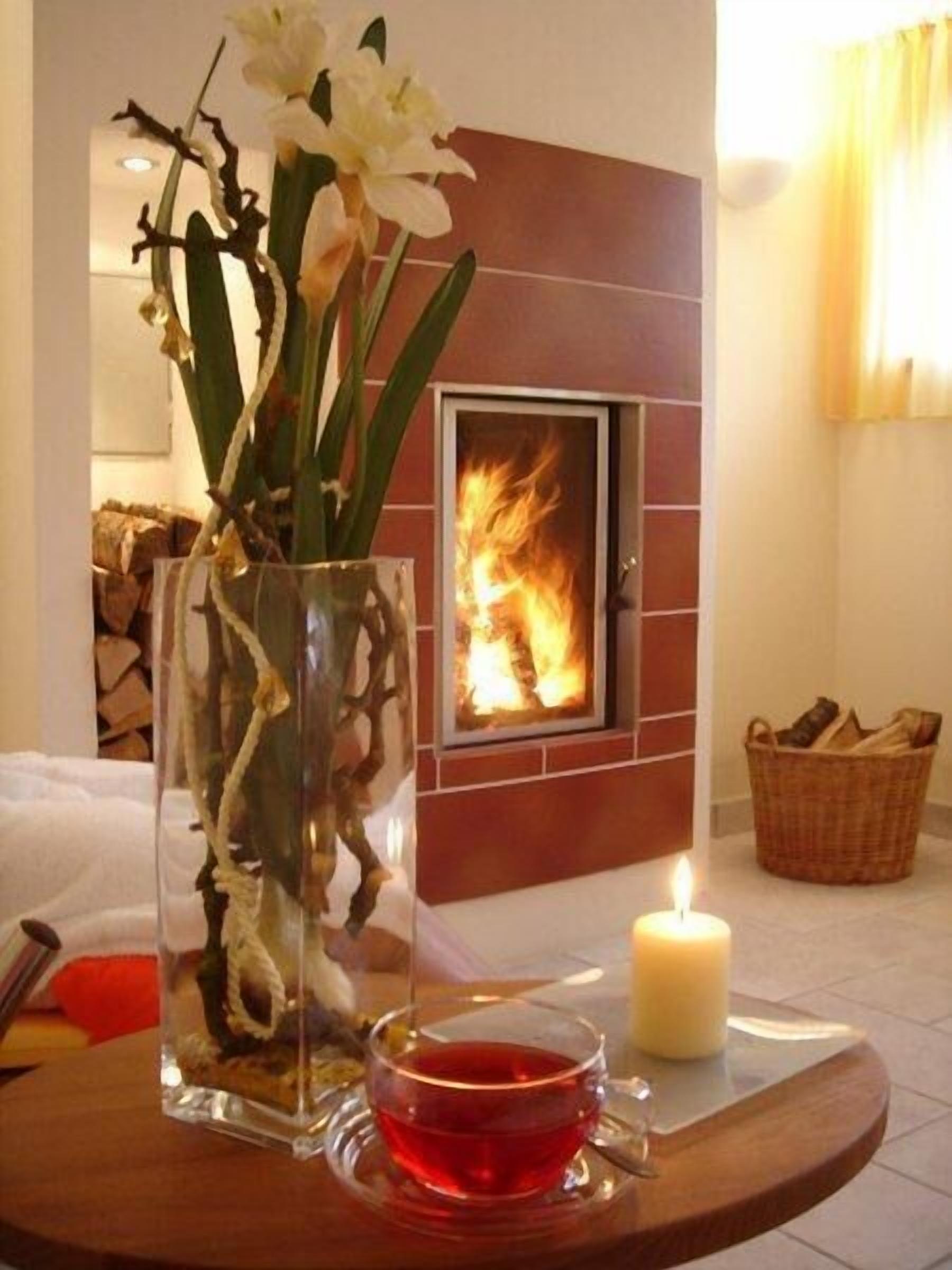 A room with a fireplace, a vase with flowers, a candle, and a table with a glass of red liquid.