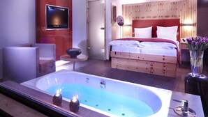 A modern hotel room with a large bathtub, a bed with white linens, a small round table, and a TV mounted on the wall. - Alpen Adria Hotel & Spa (Hermagor-Pressegger See)