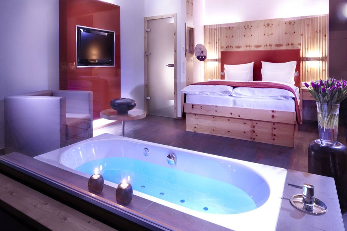 Una habitación de hotel moderna con una bañera grande, una cama con ropa blanca, una mesita redonda y un televisor montado en la pared.