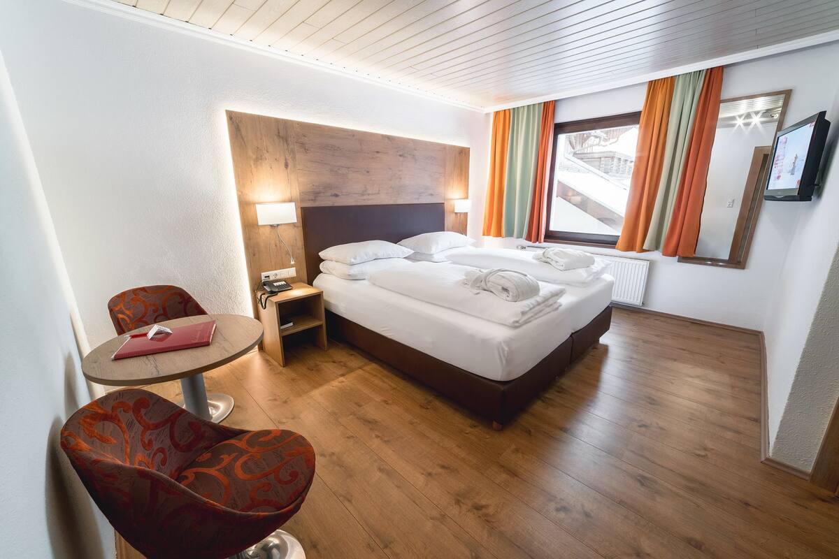 Habitación de hotel con piso de madera, una cama con ropa de cama blanca, una mesita con una carpeta roja, una silla y un televisor en la pared.
