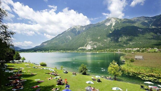 A lakeside view with a grassy area, tents, and a mountain backdrop. - Alpen Adria Hotel & Spa (Hermagor-Pressegger See)